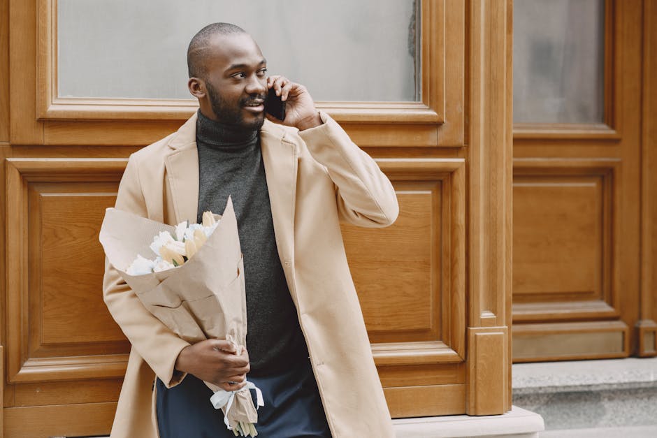 A young man with a dark beard and short hair is standing outdoors in front of a wooden paneled wall, holding a bouquet of fresh flowers wrapped in brown paper. The bouquet contains white roses and oth