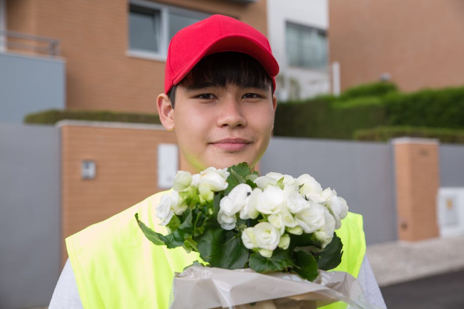 A young male flower delivery courier with light skin is standing outdoors in front of modern residential buildings, holding a bouquet of fresh white flowers with green foliage. He is wearing a bright 